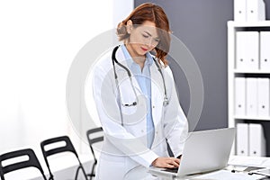 Happy doctor woman at work. Portrait of female physician using laptop computer while standing near reception desk at