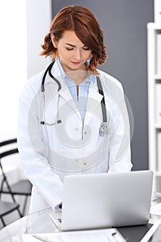 Happy doctor woman at work. Portrait of female physician using laptop computer while standing near reception desk at