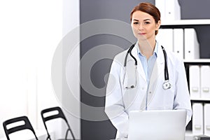 Happy doctor woman at work. Portrait of female physician using laptop computer while standing near reception desk at