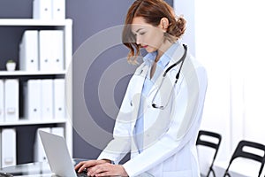 Happy doctor woman at work. Portrait of female physician using laptop computer while standing near reception desk at