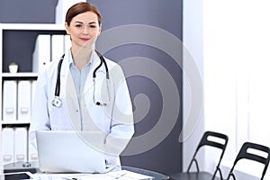 Happy doctor woman at work. Portrait of female physician using laptop computer while standing near reception desk at