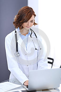 Happy doctor woman at work. Portrait of female physician using laptop computer while standing near reception desk at