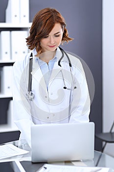 Happy doctor woman at work. Portrait of female physician using laptop computer while standing near reception desk at