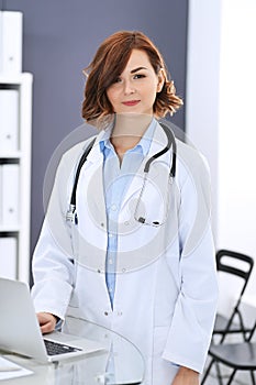 Happy doctor woman at work. Portrait of female physician using laptop computer while standing near reception desk at