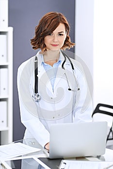 Happy doctor woman at work. Portrait of female physician using laptop computer while standing near reception desk at