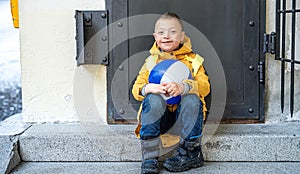 Happy down syndrome boy with ball outdoors sitting in front of door, looking at camera.