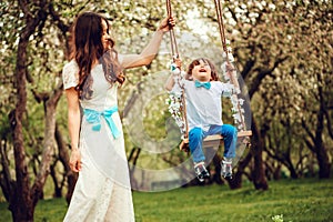 Happy dressy mother and toddler child son having fun on swing in spring or summer park