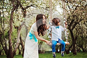 Happy dressy mother and toddler child son having fun on swing in spring or summer park, wearing bow tie and long lacy dress for bi