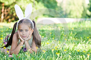 Happy Easter: adorable little girl wearing bunny ears lying on a meadow, leaning her face on her hands, outdoors