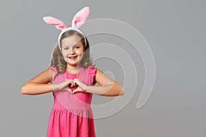 Happy easter. Cheerful little girl in a pink dress with polka dots on a gray background. A child in a rabbit costume shows a heart
