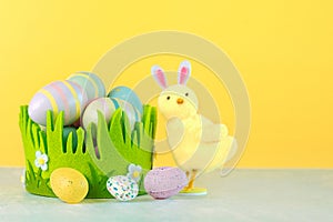 Happy Easter. Colored easter painted eggs in a green basket next to a chick with rabbit ears