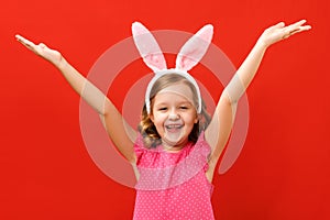 Happy easter. Cute little girl in bunny ears raised her hands up. Close-up baby on a colored red background