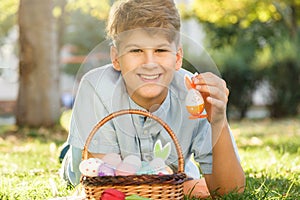 Happy Easter! Cute smiling boy teenager in blue shirt holds basket with handmade colored eggs on grass in spring park.