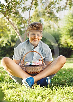 Happy Easter! Cute smiling boy teenager in blue shirt holds basket with handmade colored eggs on grass in spring park.