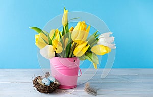 Happy Easter. Decorative easter eggs in bird nest and bouquet of yellow tulips in bucket on blue background. Concept: beautiful