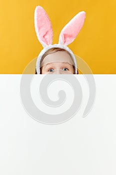 Happy easter. Funny little girl with bunny ears is hiding because of a blank banner. The child preys on eggs