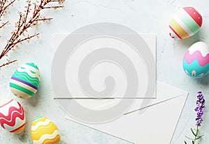 Happy Easter greeting card mockup, blank paper with pastel color eggs, envelope, flowers and wooden table. Empty card design