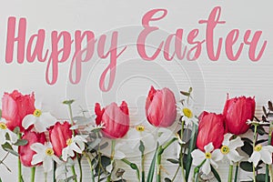 Happy Easter greeting card. Happy Easter text sign handwritten on  beautiful red tulips, daffodils, daisy flowers on rustic white