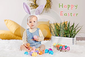 Happy Easter. Greeting holiday card with text. Caucasian baby girl with purple bunny ears sitting on a fluffy white rag carpet.