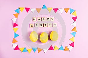 Happy easter inscription in wooden letters with yellow painted eggs in a frame made of multicolored garland of felt on a pink