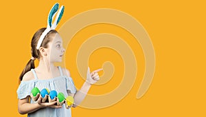 Happy Easter, kids. A girl in bunny ears on her head holds a tray with eggs of blue and yellow color, the emotion of surprise on