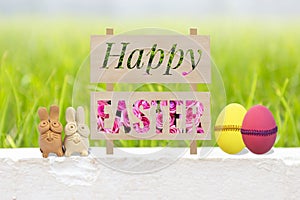 Happy Easter sign with clay rabbit and easter eggs over blurred green grass background