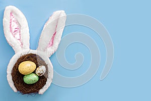 Happy easter and spring meme concept with adorable bunny ears on a bird nest with colorful eggs against a baby blue background