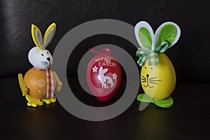 Two easter rabbit figure made of plastic egg, dark background. Green-white and and orange checkered pattern ribbon bow, Yellow-ora