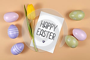 `Happy Easter` text card surrounded by colorful easter eggs and single yellow tulip spring flower on beige colored background, fla