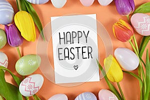 `Happy Easter` text card surrounded by colorful easter eggs and tulip spring flowers on orange background, flat lay