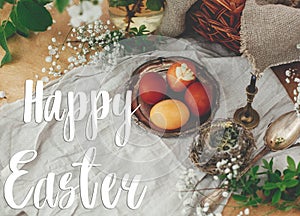 Happy Easter text. Easter greetings lettering. Modern Easter eggs on vintage plate on rustic table with spring flowers,candle,