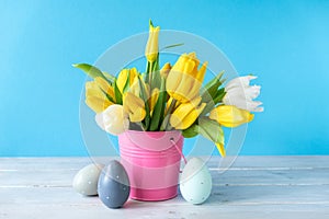 Happy Easter. Wooden decorative eggs and bouquet of yellow tulips in bucket on blue background. Concept: beautiful greeting easter