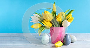 Happy Easter. Wooden decorative eggs and bouquet of yellow tulips in bucket on blue background. Concept: beautiful greeting easter