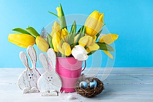 Happy Easter. Wooden decorative two funny  bunnies, eggs in bird nest and bouquet of yellow tulips in bucket on blue background.