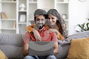 Happy eastern couple websurfing on laptop together while relaxing at home