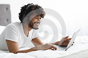 Happy eastern guy enjoying weekend in bed, using laptop