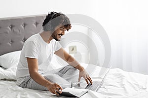 Happy eastern guy studying in bed, using laptop