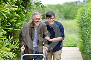 Happy elder Asian man using walker while walking for exercise around the garden with his son taking care of him at the retirement