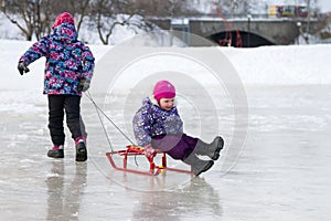 Happy elder girl pulling her young sister on a sled on the ice in snowy winter park