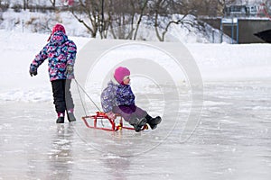 Happy elder sister pulling her young sister on a sled on the ice in snowy winter park
