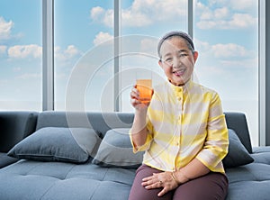 Happy elderly Asian woman sitting on sofa and hold glass of orange juice in living room at home