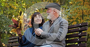 Happy elderly couple enjoy outdoor activities sitting on a bench. Wife holds a bouquet of autumn leaves. The husband