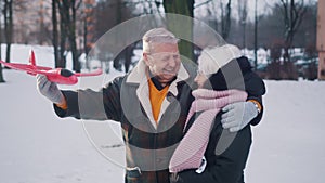 Happy elderly couple hugging and playing with airplane toy in the park in winter. Slow motion