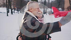 Happy elderly couple hugging and playing with airplane toy in the park in winter. Slow motion