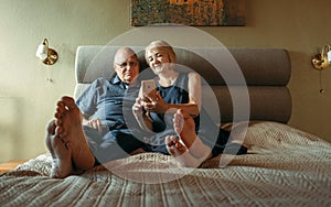 Happy elderly couple of pensioners, looking at the screen of a mobile phone, lie on the bed