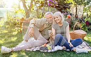 Happy elderly couple relaxing in garden and using smartphone, sitting on blanket on grass, resting outdoors, copy space