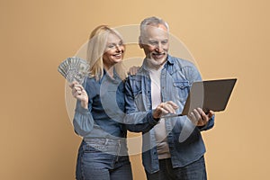 Happy elderly couple spouses using computer laptop, holding cash