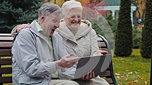 Happy elderly grandparents couple family senior man husband and middle-aged woman wife sit on bench outdoors use laptop