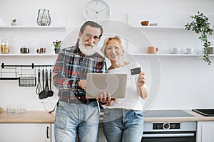 Happy elderly husband and wife holding laptop and credit card paying for food.