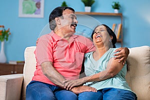 Happy elderly Indian senior couple laughing together while sitting closely at home - concept of companionship, affection